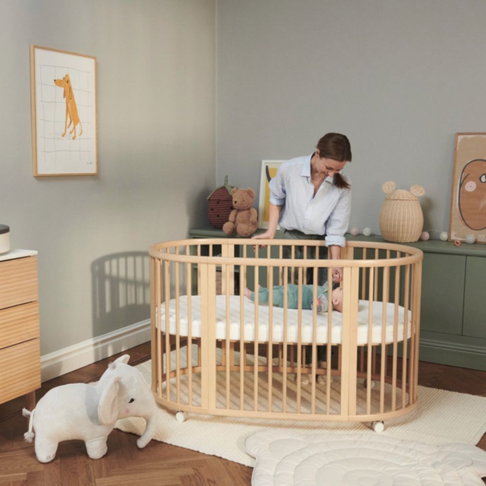 Woman tending to a baby in a wooden crib in a nursery. This is Stokke Sleepi Bed Natural - Adjustable & Mobile Scandinavian Design Crib with Lockable Wheels