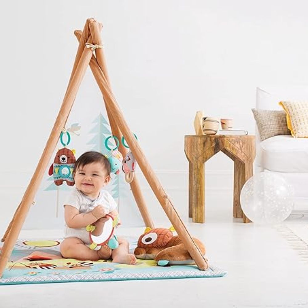 Baby sitting on a play mat with a wooden teepee and toys in a room.