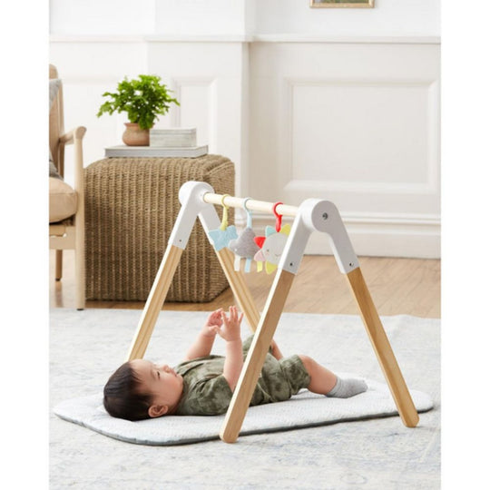 Baby playing with Skip Hop Comfy Plush Wooden Activity Gym on a white rug in a room.