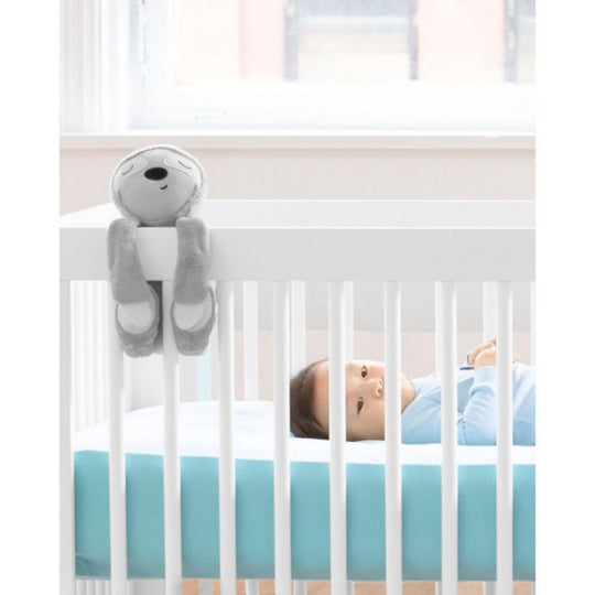 Baby lying in a crib with a gray sloth hanging over the side.