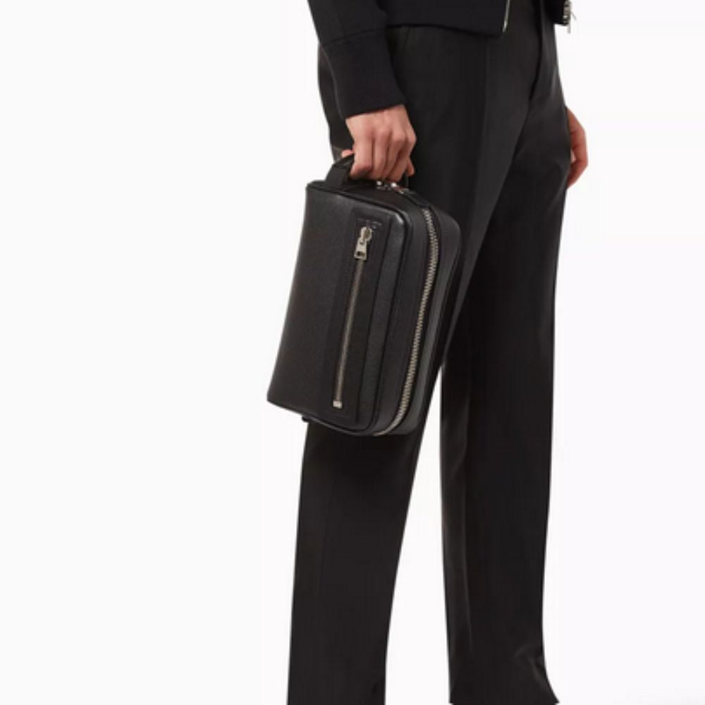 A person in black attire, including a dark jacket and trousers, holds a black, rectangular Tom Ford wash bag in their right hand. The wash bag features a textured leather surface and a prominent silver-tone zipper running along its side.