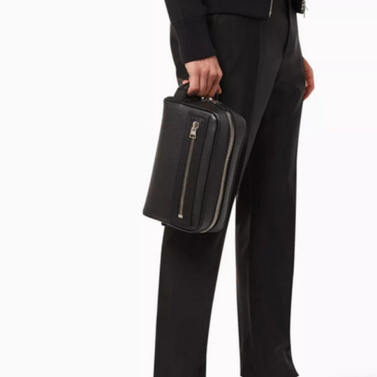 A person in black attire, including a dark jacket and trousers, holds a black, rectangular Tom Ford wash bag in their right hand. The wash bag features a textured leather surface and a prominent silver-tone zipper running along its side.