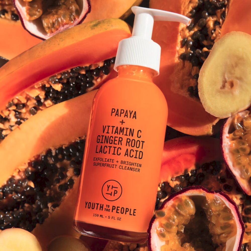 A vibrant orange bottle of YOUTH TO THE PEOPLE Superfruit Gentle Exfoliating Cleanser with a white pump top, is positioned amidst a scattering of sliced papayas and passion fruits. The bottle's label highlights key ingredients like "PAPAYA + VITAMIN C GINGER ROOT LACTIC ACID" and states "EXFOLIATE + BRIGHTEN SUPERFRUIT CLEANSER"