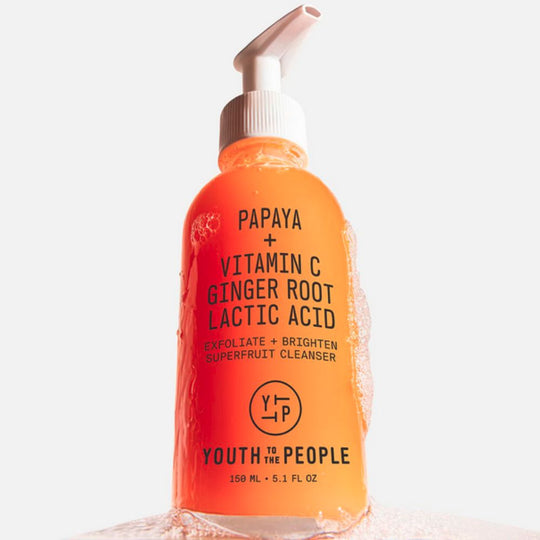 A brightly lit, close-up shot of the YOUTH TO THE PEOPLE Superfruit Gentle Exfoliating Cleanser. The orange bottle with a white pump has foam or bubbles clinging to its side, suggesting its use as a cleanser. The label is clearly visible, listing "PAPAYA + VITAMIN C GINGER ROOT LACTIC ACID" and describing it as an "EXFOLIATE + BRIGHTEN SUPERFRUIT CLEANSER". The product is on a light, possibly foamy or liquid, surface
