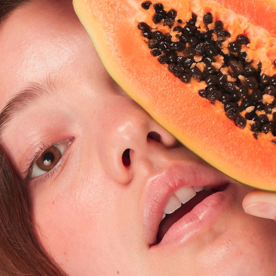 A close-up shot shows a woman's face partially obscured by a slice of papaya. The papaya covers her eye and cheek, highlighting the fruit's vibrant orange color and dark seeds.
