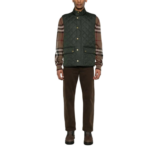 Person wearing a green quilted vest over a plaid shirt with brown pants on a white background