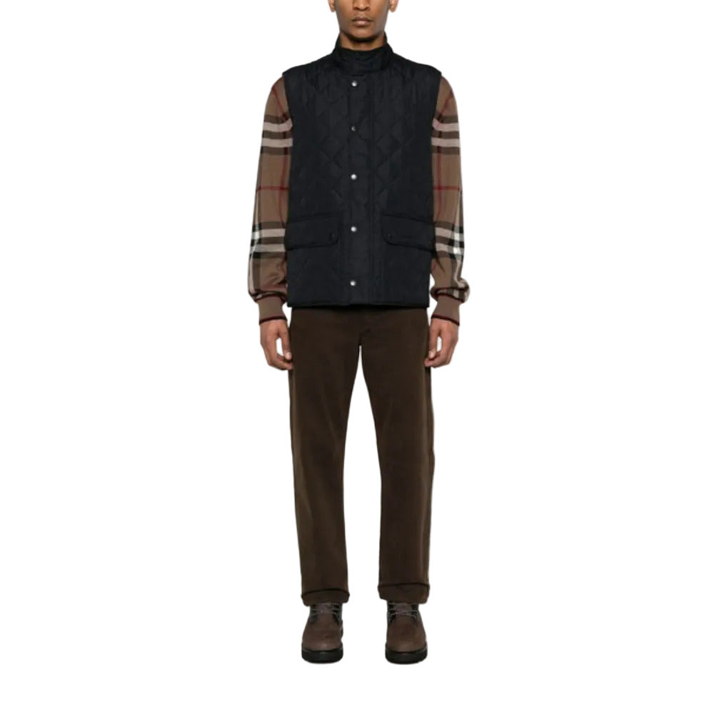 Person wearing a dark quilted vest over a plaid shirt with brown pants on a white background