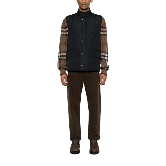 Person wearing a dark quilted vest over a plaid shirt with brown pants on a white background