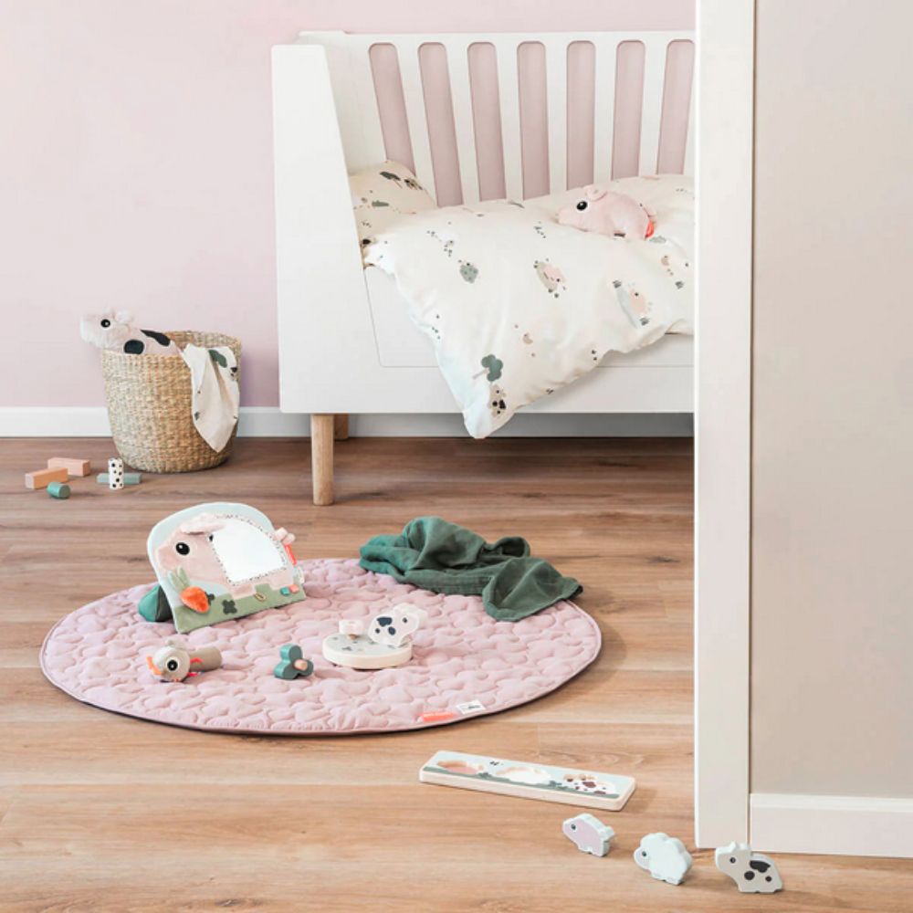 Children's play area with a Done by Deer pink mat, toys, and a crib in a room.