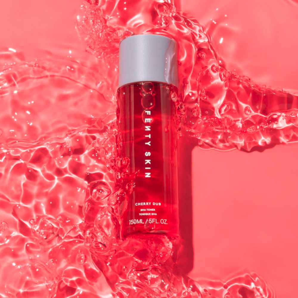 A Fenty Skin Cherry Dub BHA Toner bottle stands upright in a pool of vibrant red water with ripples and splashes extending outwards. The bottle is clear with a silver cap, and the "FENTY SKIN" logo is prominently displayed vertically on its front. Below the logo, "CHERRY DUB," "BHA TONER," and "150ML / 5FL.OZ" are visible. 