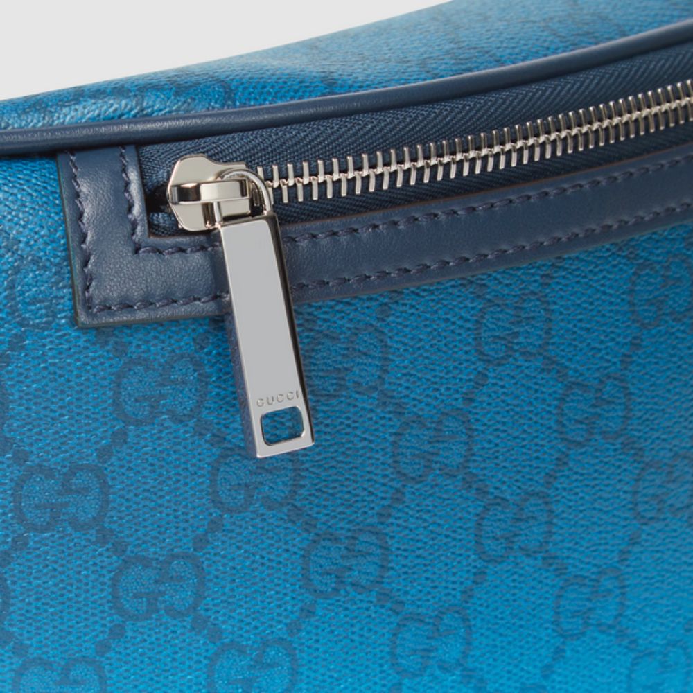 A close-up shot of a blue Gucci belt bag, showcasing its gradient color effect that transitions from dark to light blue. The bag features the repeating GG monogram pattern and a prominent silver-toned zipper with a "GUCCI" engraved pull tab.