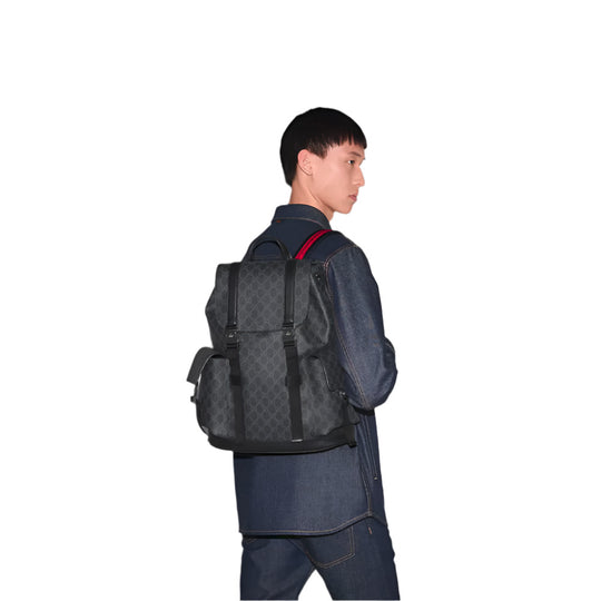 A full-body shot of a person with short dark hair, wearing a dark blue denim jacket and pants, facing right and looking over their left shoulder. They are carrying a GUCCI Chroma Large Backpack on their back, which is black with the GG monogram pattern. The backpack has black straps and silver-toned hardware, with a subtle red stripe detail visible on the left shoulder strap.