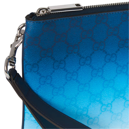 An extreme close-up of a blue Gucci pouch, showing its gradient color effect and the GG monogram pattern. A silver-toned D-ring and a dark blue leather wrist strap are visible on the left side, along with part of a zipper.