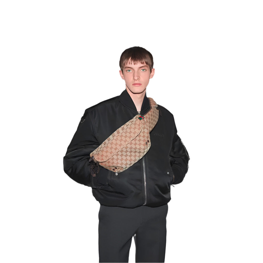 A fair-skinned person with short, light brown hair and blue eyes stands against a plain white background, wearing a black bomber jacket, dark trousers, and a large GUCCI Beige and dark brown GG Canvas Large Belt Bag slung across their chest. The bag features the brand's signature beige and brown monogram pattern with the red and green Web stripe visible near the top. 