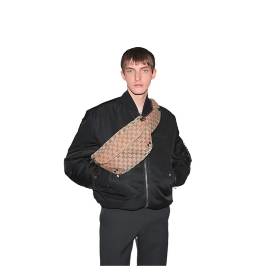 A fair-skinned person with short, light brown hair and blue eyes stands against a plain white background, wearing a black bomber jacket, dark trousers, and a large GUCCI Beige and dark brown GG Canvas Large Belt Bag slung across their chest. The bag features the brand's signature beige and brown monogram pattern with the red and green Web stripe visible near the top. 