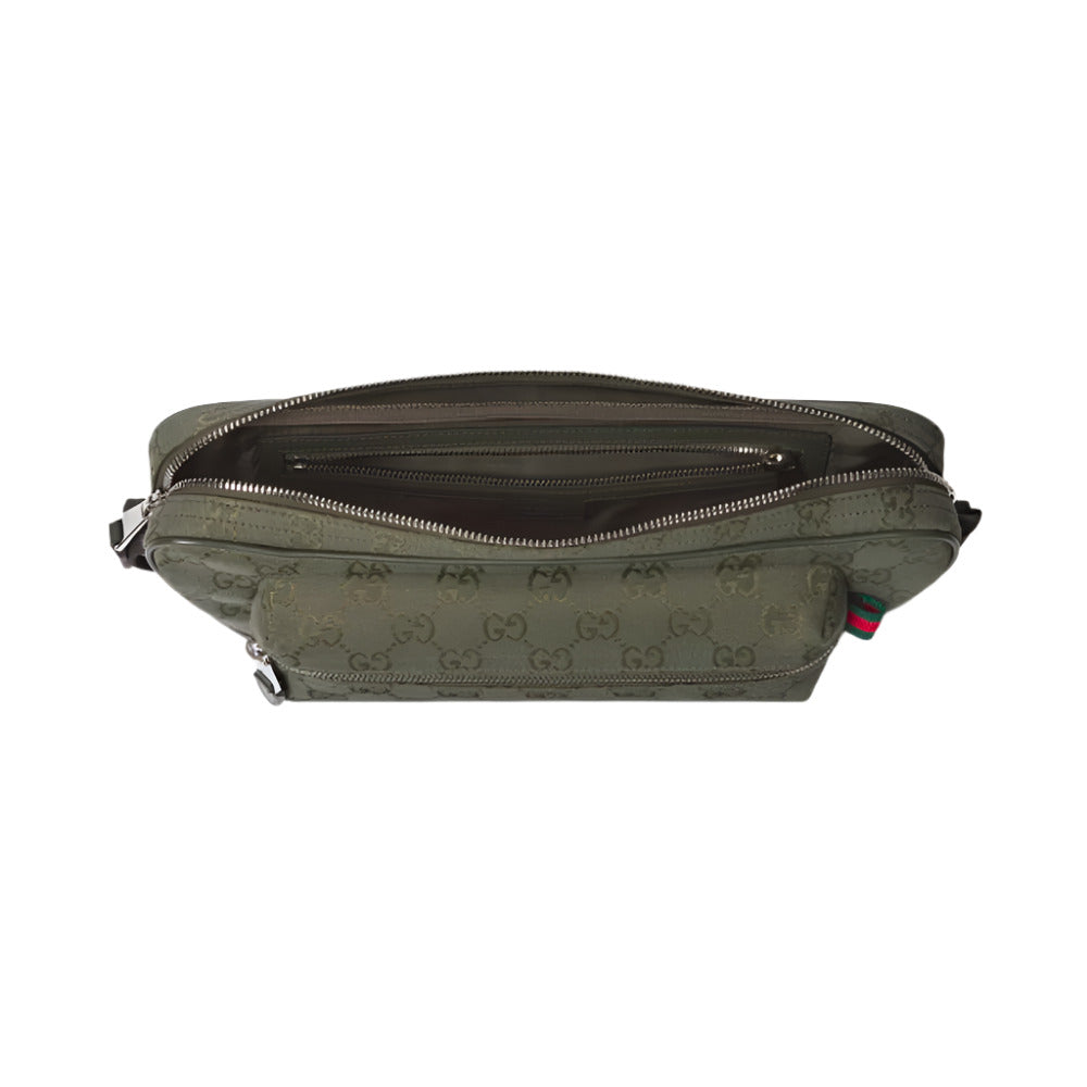 An overhead shot of the GUCCI GG Canvas Large Slingbag, showing the top open. The bag is made of forest green GG canvas with the interlocking GG monogram subtly debossed or printed throughout. The interior is lined with a darker green nylon, and several open pockets and one zippered pocket are visible. On the exterior, near the front, there are two zippered pockets. On the right side, a small tag with the iconic green and red Web detail is visible.