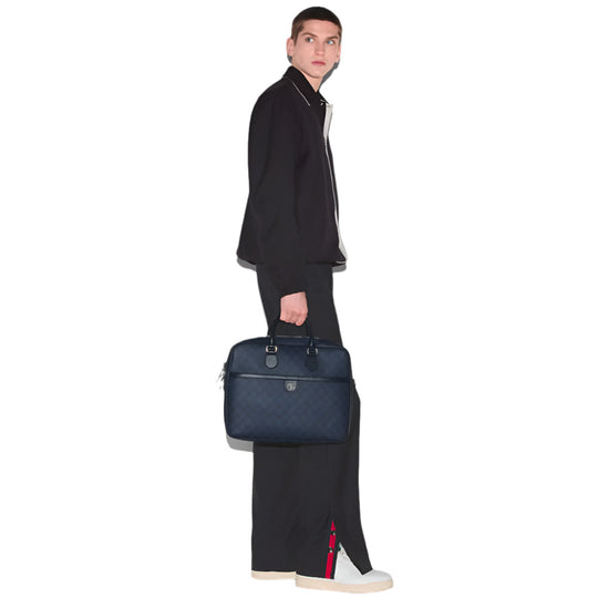 A man in a dark blue jacket and trousers with a red and green stripe holds a dark blue briefcase with a GG logo, against a white background.