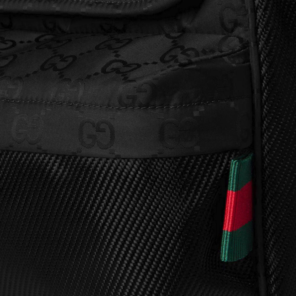 A close-up of the black GUCCI Nexus Medium Crossbody Bag, showing the tonal GG monogram on the fabric above a textured panel, and a small red and green Web stripe tag on the side.