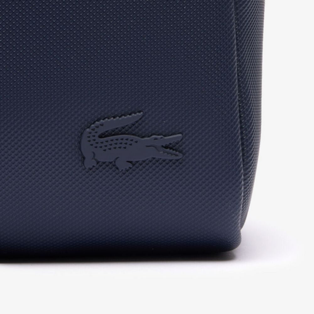 A close-up of a navy blue Lacoste toiletry bag, featuring the brand's iconic tone-on-tone crocodile logo debossed on its textured, piqué-effect canvas surface.