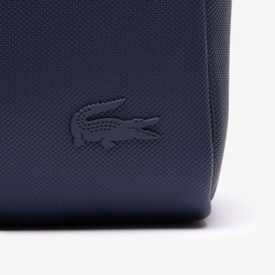 A close-up of a navy blue Lacoste toiletry bag, featuring the brand's iconic tone-on-tone crocodile logo debossed on its textured, piqué-effect canvas surface.