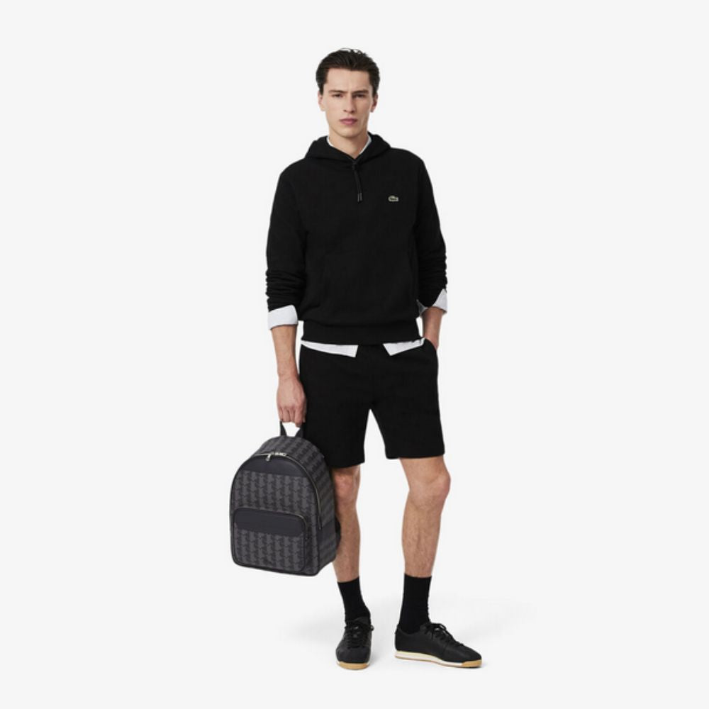 A person stands facing forward, holding a Lacoste The Blend Backpack in their left hand. They are dressed in a black hoodie with a white shirt underneath, black shorts, black socks, and black sneakers with tan soles. The backpack features a dark grey and black monogram pattern with black leather accents. The background is plain white.