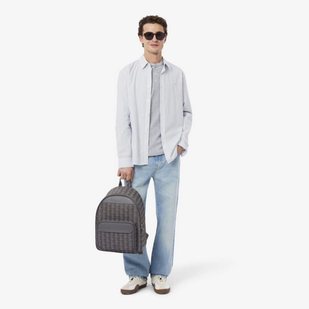 A light-skinned person stands facing the viewer, wearing sunglasses, a light-colored button-up shirt over a grey t-shirt, light blue jeans, and light-colored sneakers with tan soles. They hold a Lacoste The Blend Backpack in their left hand, which features a grey and black monogram pattern with solid grey accents and a front zip pocket. 