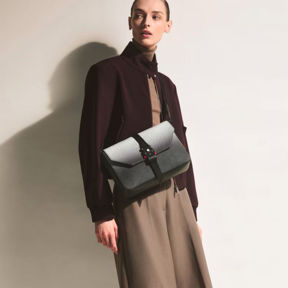 A person wearing a dark red jacket and tan pants stands with a black and white gradient Montblanc Extreme 3.0 Messenger with M LOCK Buckle & Adjustable Strap slung over their shoulder. 