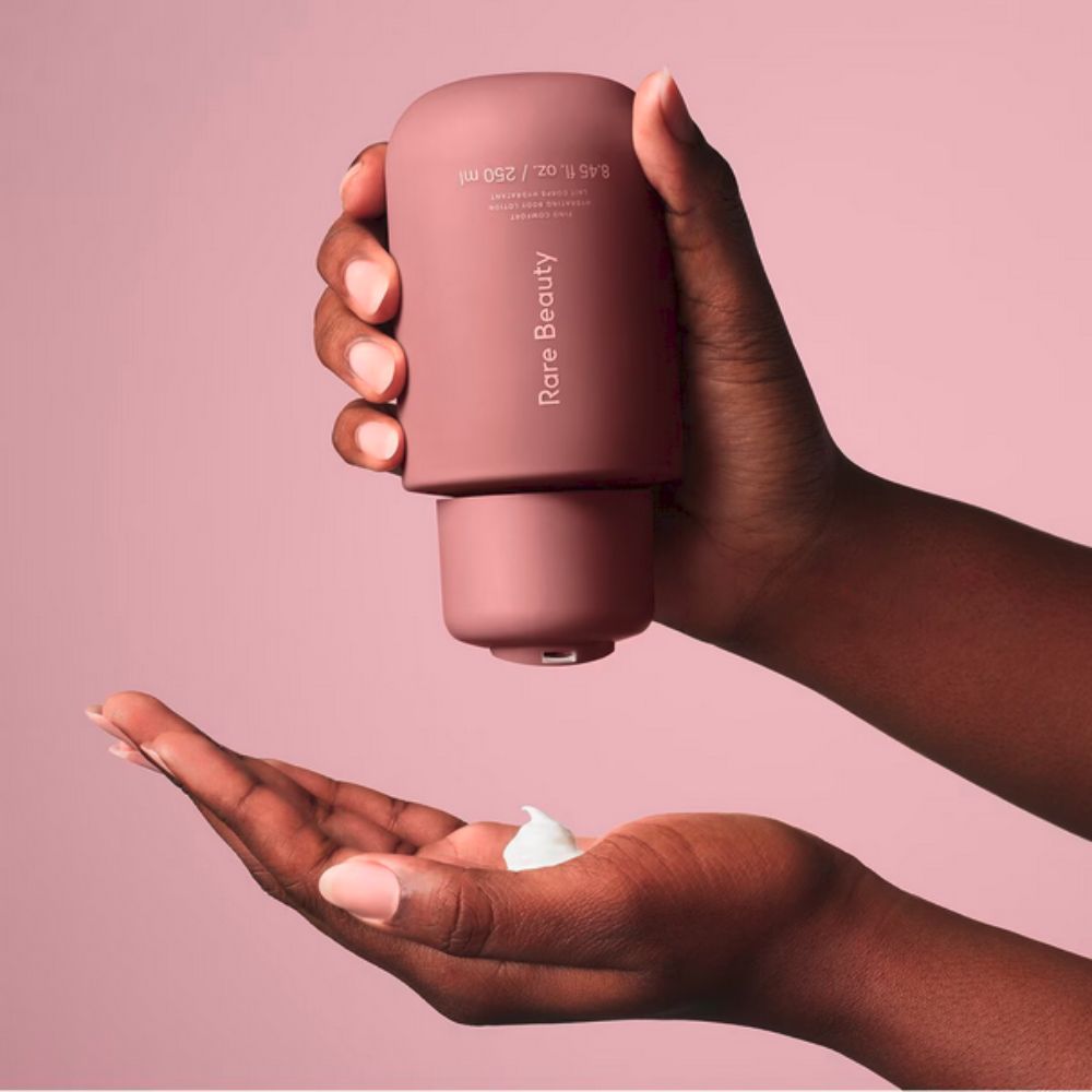 A pair of hands, with short, neat nails, hold a rose-pink, minimalist Rare Beauty Find Comfort Hydrating Body Lotion bottle. The upper hand squeezes the bottle, dispensing a dollop of white lotion into the open palm of the lower hand. 