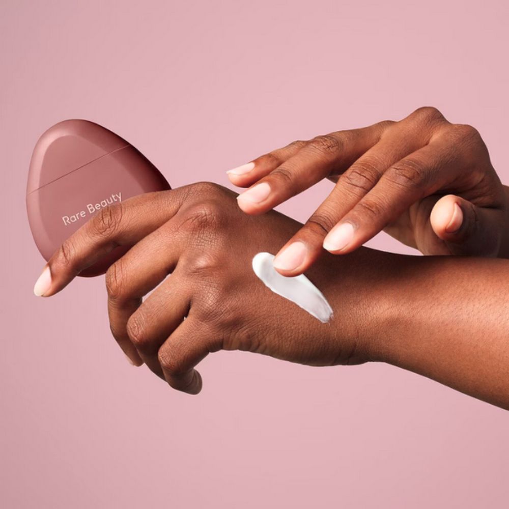 A pair of hands with dark skin tones are shown applying a dollop of white cream to the back of one hand. The cream is being spread by the fingers of the other hand. In the background to the left, the bottom portion of a rose-colored, ergonomically shaped Rare Beauty hand cream bottle is visible. 
