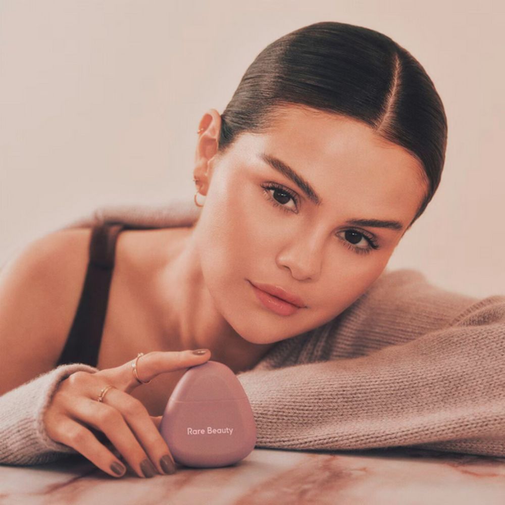 Selena Gomez, with a neutral expression and light skin tone, rests their chin on their left arm, which is clad in a light grey knitted sweater. Their right hand, adorned with several rings and painted fingernails, gently holds a smooth, egg-shaped, light purple Rare Beauty product, likely a hand cream. 