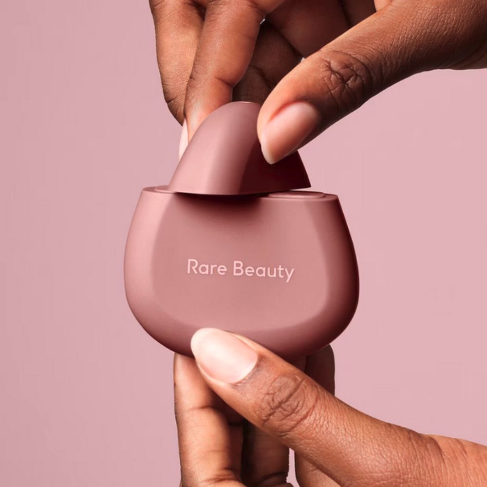 Two hands with dark skin tones are shown opening a light pink, egg-shaped Rare Beauty hand cream bottle. The top of the bottle is being twisted off to reveal the opening, while the base is held steady. 