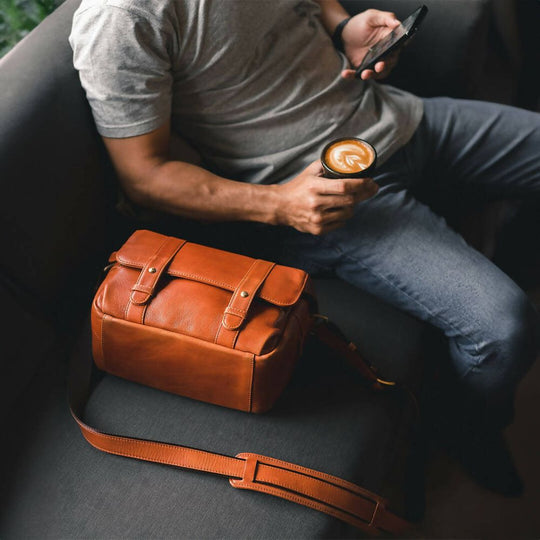 Person sitting on a couch holding a cup of coffee with a brown leather bag next to them.
