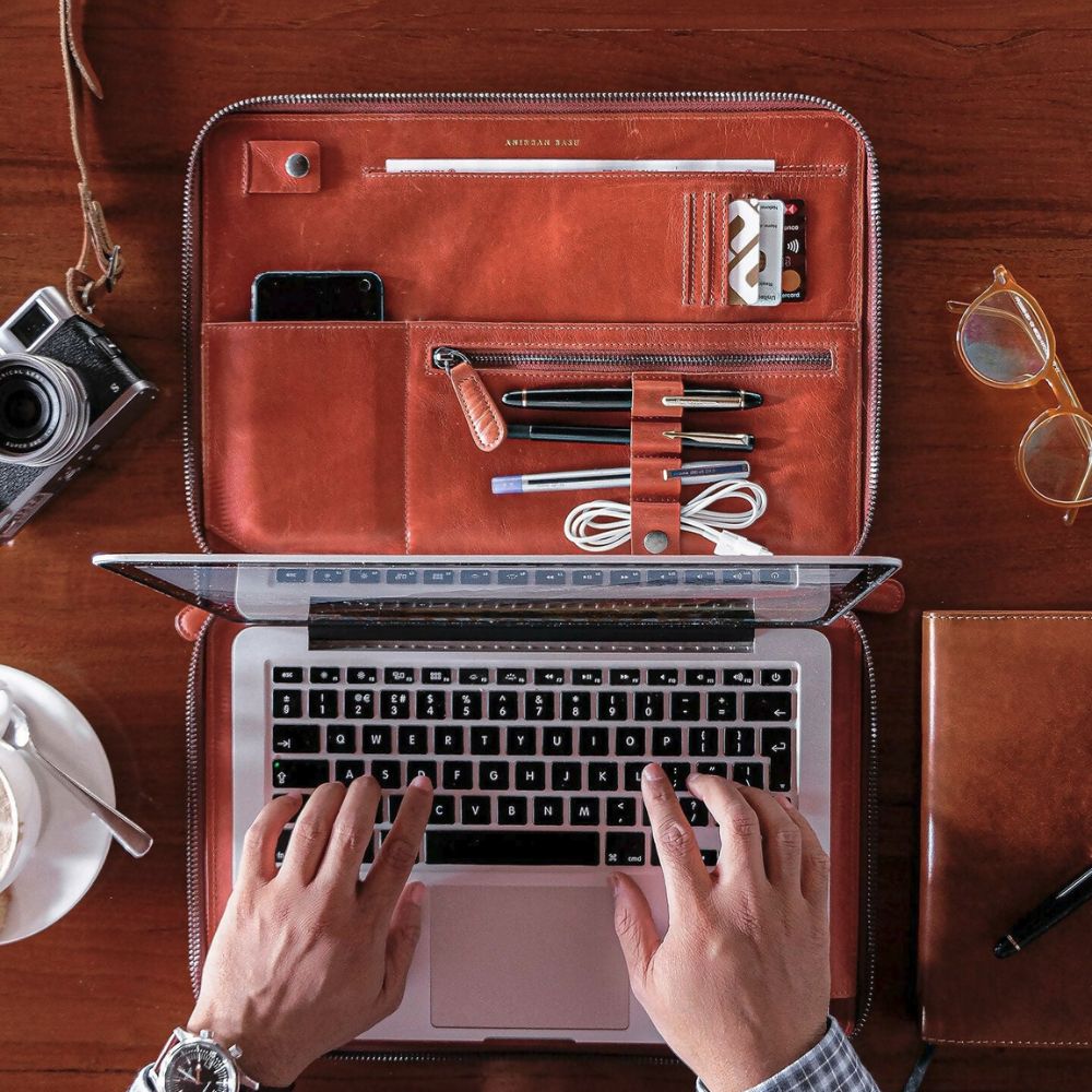 Person using a laptop with a leather case on a wooden desk, surrounded by items like a camera, sunglasses, and a notebook.