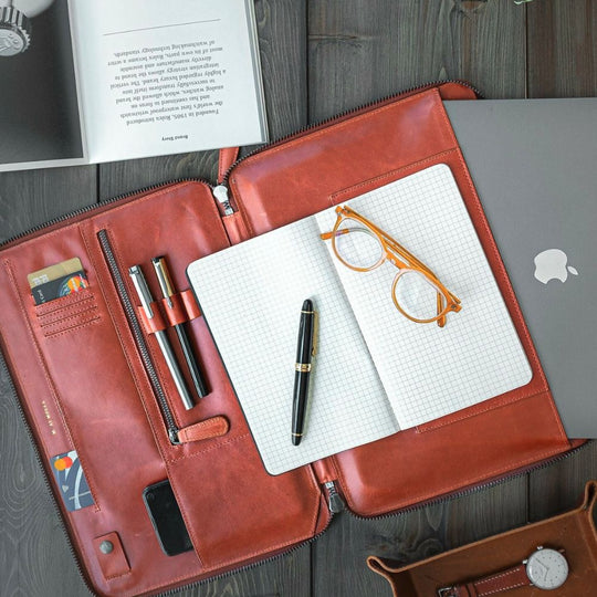 Brown leather portfolio with notebook, pen, glasses, and phone on a wooden surface.