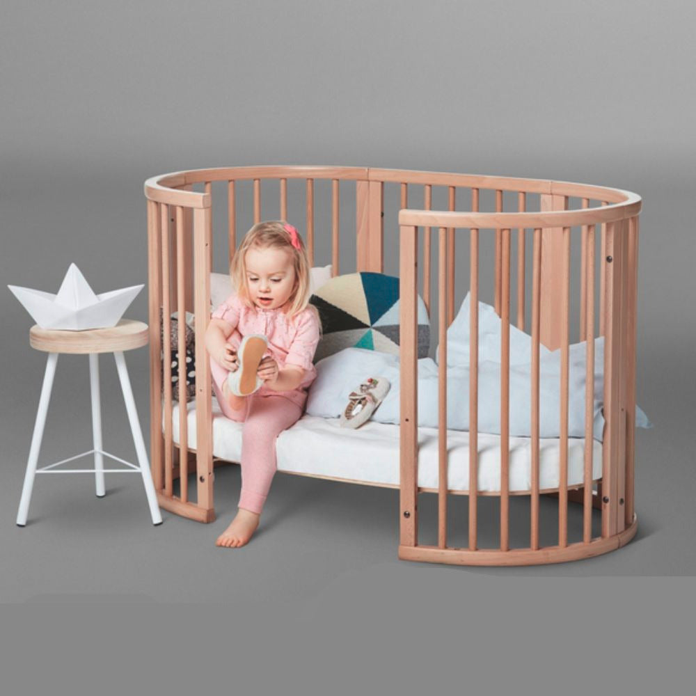 Child sitting in a wooden crib with a removable panel, gray background. The Crib is a Stokke Sleepi Bed Natural - Adjustable & Mobile Scandinavian Design Crib with Lockable Wheels