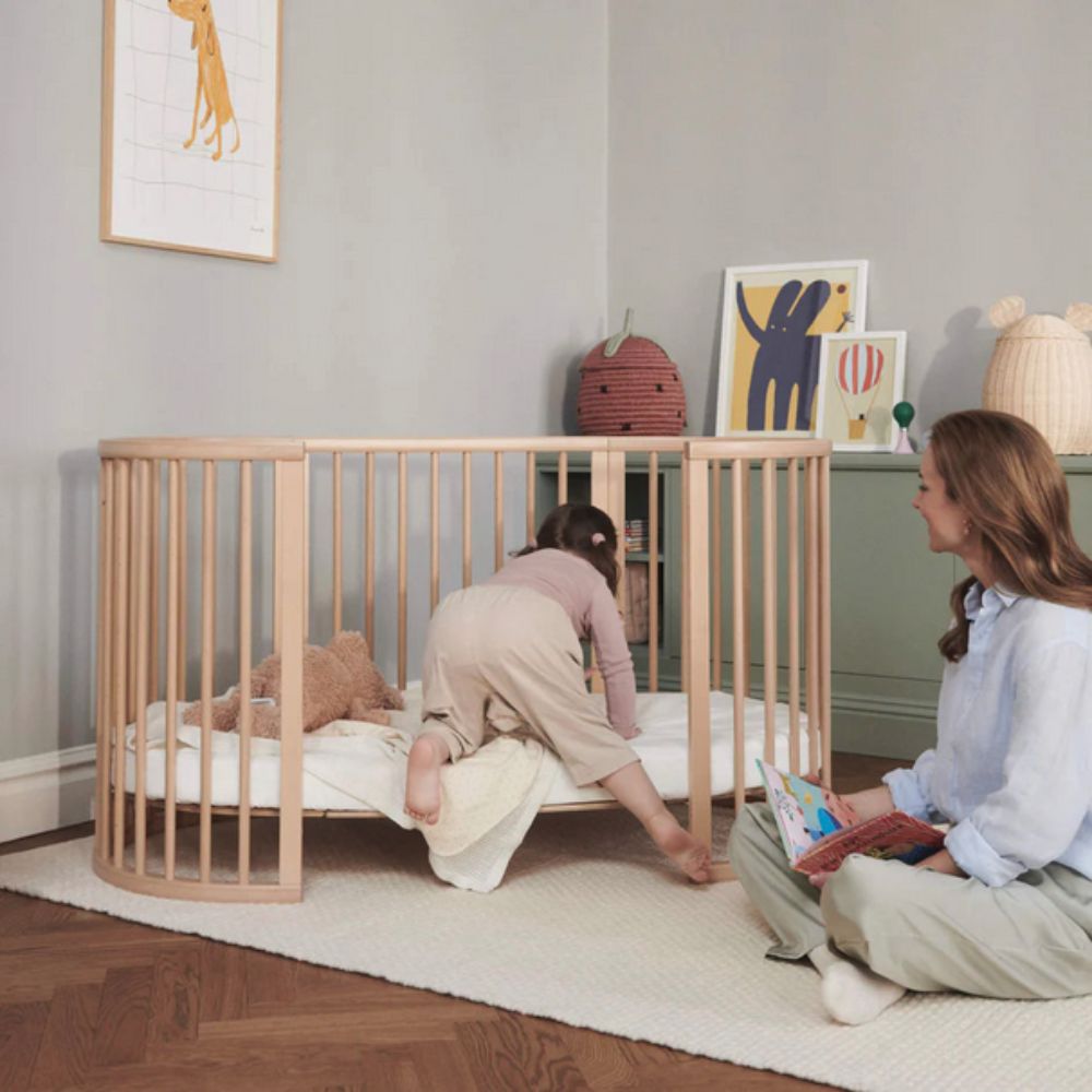 Woman reading to a child in a crib in a cozy room. The Crib is a Stokke Sleepi Bed Natural - Adjustable & Mobile Scandinavian Design Crib with Lockable Wheels