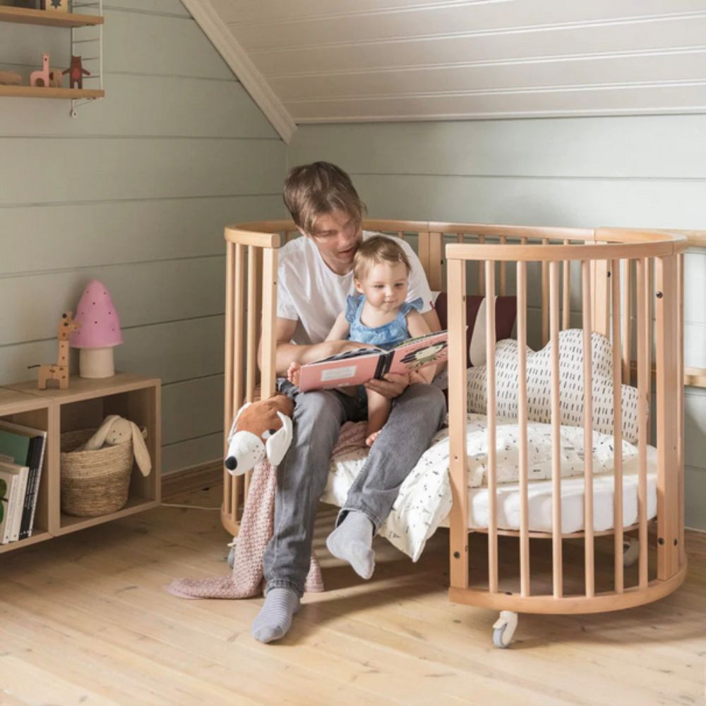 Man reading a book to a child in a wooden crib in a cozy room. This is a Stokke Sleepi Bed Natural - Adjustable & Mobile Scandinavian Design Crib with Lockable Wheels