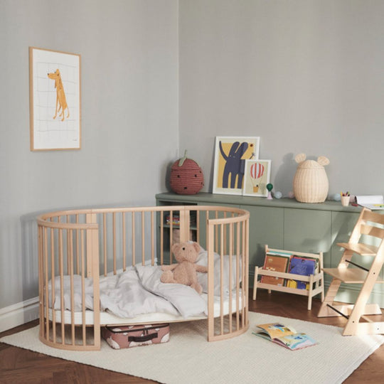 Children's room with a round wooden crib, teddy bear, and books. The crib is a Stokke Sleepi Bed Natural - Adjustable & Mobile Scandinavian Design Crib with Lockable Wheels