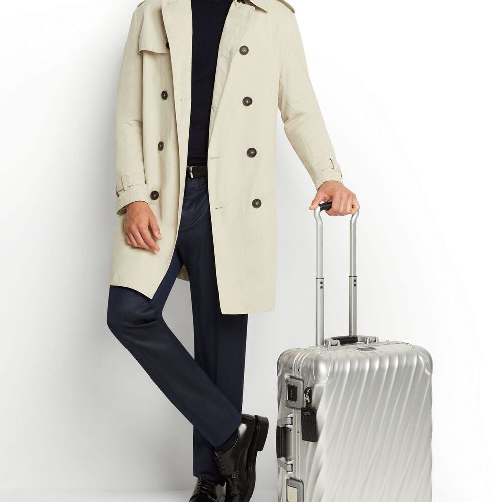Person wearing a beige trench coat holding a silver suitcase on a white background