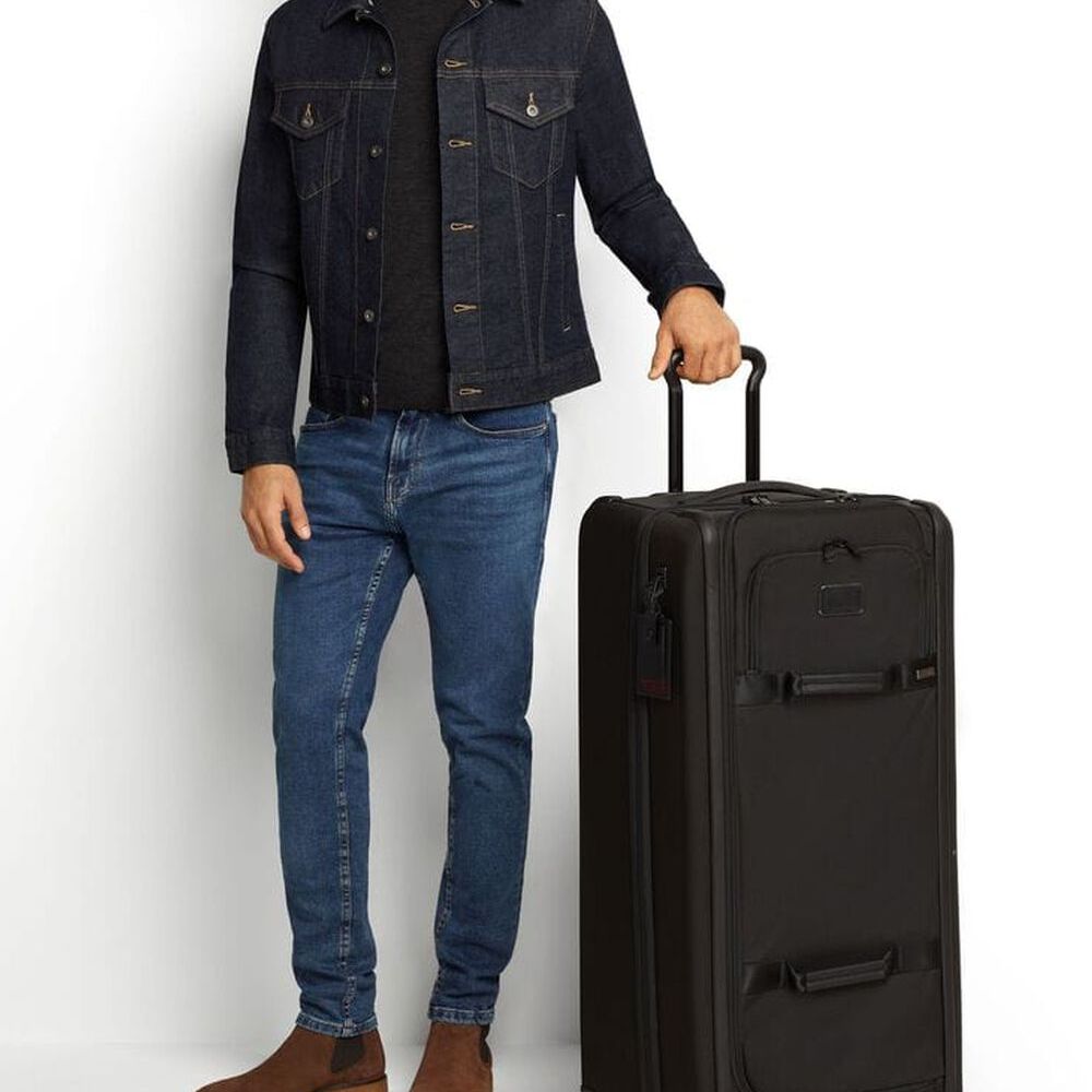 A model standing besides TUMI Versatile Alpha 3 Tall Wheeled Duffel: Model Number 117169-1041, holding its extended handle