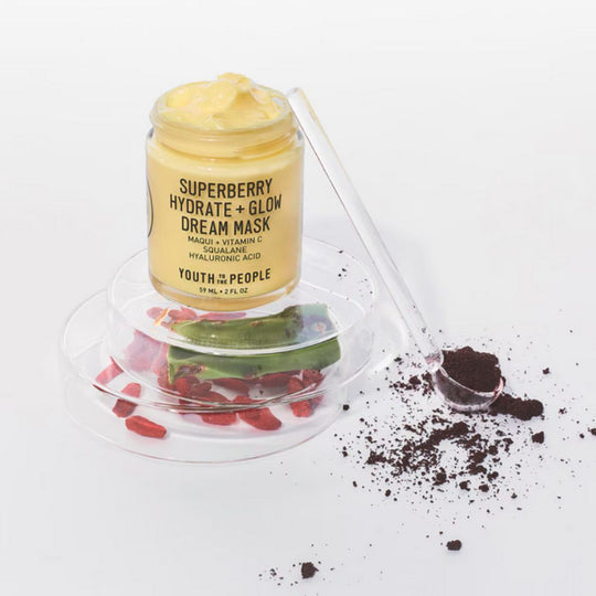A studio shot of the YOUTH To The PEOPLE Superberry Hydrate + Glow Dream Mask, featuring a glass jar with a yellow cream inside. The jar sits on a clear, layered platform with scattered goji berries, a sliced aloe leaf, and a spoonful of dark powder. 